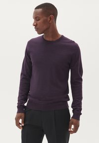 Ljubičasti pulover s okruglim izrezom i dugim rukavima, s rebrastim manžetama i donjim rubom, izrađen od mekanog materijala, uparen s crnim hlačama.