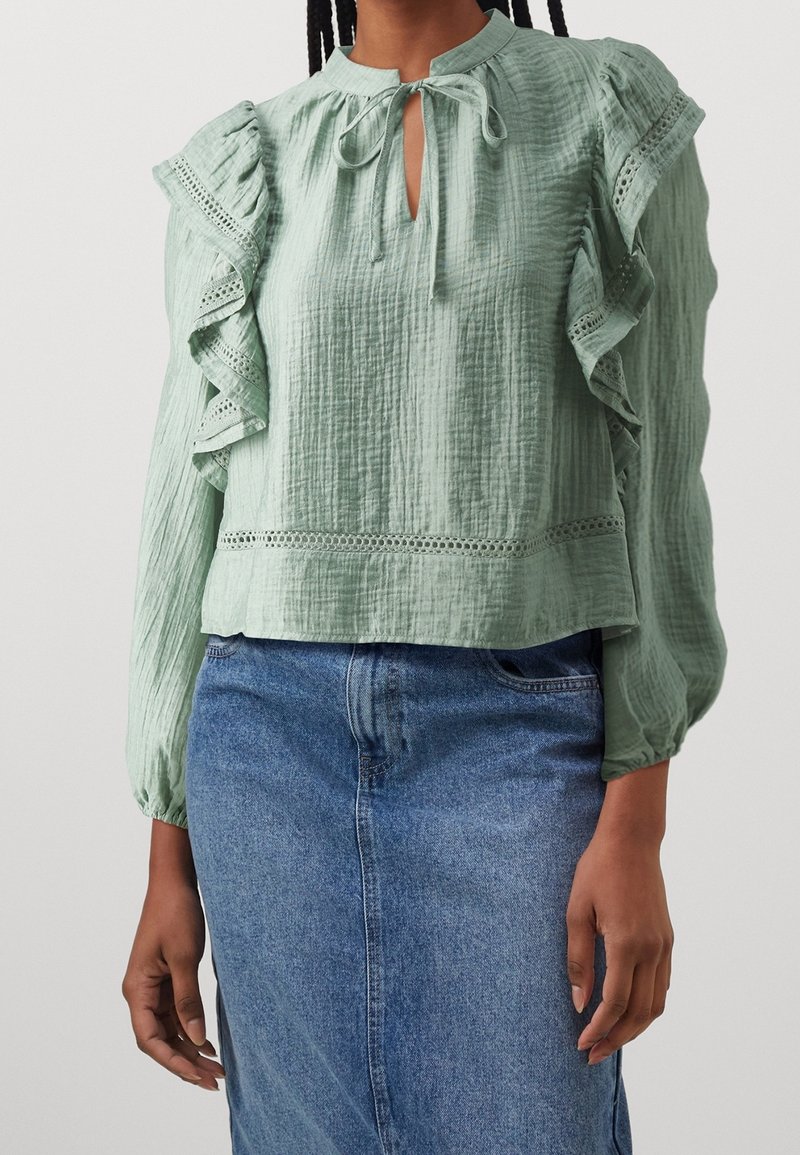 Blouse vert clair texturée avec des manches volantées, un décolleté en trou de serrure et des détails en dentelle. Portée sur une jupe en denim bleu.