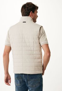 Light beige puffer vest with a high collar, quilted design, and a sleek black label at the back, paired with short sleeves and blue jeans.