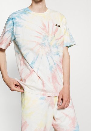 Person trägt ein pastellfarbenes Batik-T-Shirt und passende Shorts mit kleinem geflügeltem Augapfel-Design auf der oberen linken Brust.