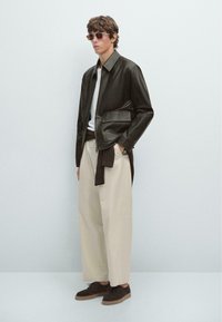 Brown leather zip-up jacket with a chest pocket, paired with loose beige trousers and dark brown suede shoes. Accessories include dark sunglasses.