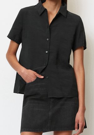 Kobieta w czarnej koszuli z krótkim rękawem zapinanej na guziki, lekko wpuszczonej w czarną spódnicę dżinsową, z lewą ręką w kieszeni.