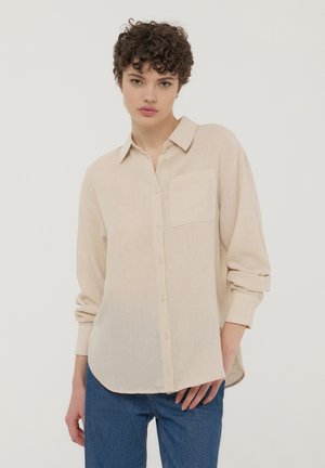 Giovane con capelli corti e ricci, che indossa una camicia beige a maniche lunghe con bottoni e jeans blu, in piedi davanti a uno sfondo bianco semplice.