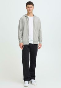 Sweat à capuche gris à fermeture éclair avec capuche, manches longues et poches avant, associé à un t-shirt blanc et un jean noir, complété par des baskets blanches.