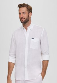 Camisa blanca de lino con cuello abotonado, mangas largas enrolladas hasta los codos, un bolsillo en el pecho y una pequeña etiqueta de marca en el bolsillo.