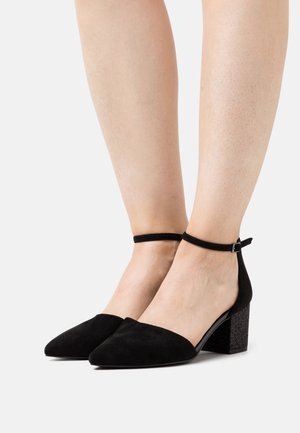 Paire de talons noirs en daim à bouts pointus avec brides à la cheville et talons carrés, portés pieds nus sur un fond blanc.