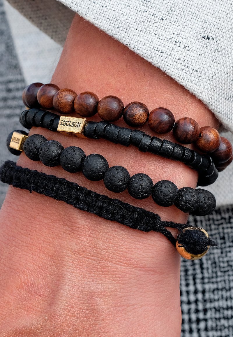 Set aus vier Armbändern mit Holzkugeln, schwarzen Lavakugeln und geflochtenem schwarzem Cord, ergänzt durch metallische Akzente und verschiedene Texturen.