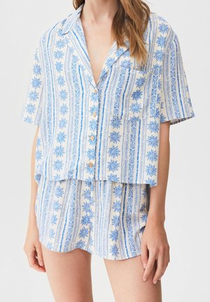Kobieta ubrana w niebiesko-białą piżamę składającą się z koszuli na guziki z krótkim rękawem w kwiaty i paski oraz pasujące krótkie spodenki.