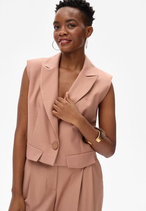 Kobieta ubrana w różową, bezrękawnikową marynarkę i dopasowane spodnie, uzupełnione kolczykami kołami i złotą bransoletką z futrzanym akcentem.