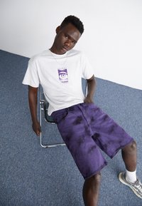 Camiseta gris con un diseño gráfico morado, combinada con pantalones cortos de tie-dye morado hasta el muslo. Sentado en una silla de metal sobre un suelo alfombrado.