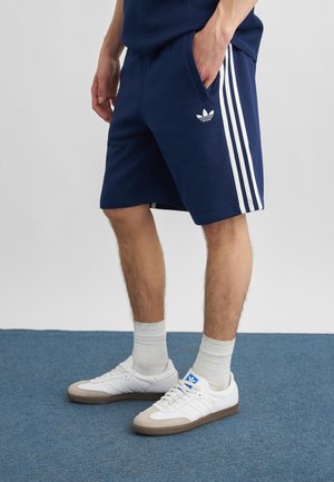 Mężczyzna w granatowych szortach Adidas z białymi paskami, białych butach sportowych Adidas i białych skarpetkach, stojący na niebieskim wykładzinowym dywanie.
