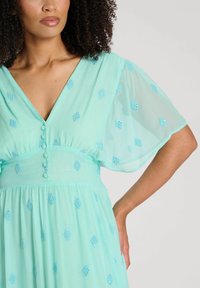 En mintgrön klänning av lätt tyg med V-ringning, korta genomskinliga ärmar och dekorativa blå broderade mönster.