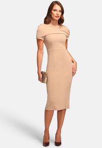 Fitted beige dress with short sleeves and draped bodice, extending below the knee. Paired with a metallic clutch and brown heels.