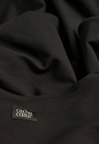 Schwarzer, dehnbarer Stoff mit glatter Textur, einer genähten Kante und einem Etikett mit der Aufschrift "cache cœur". Weiche drapierte Optik.