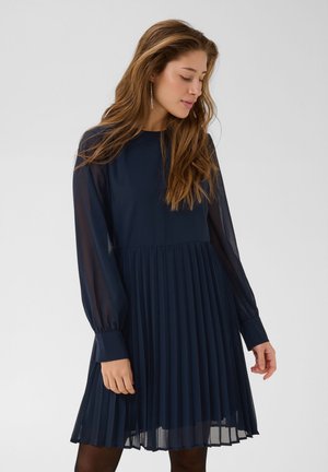 Une femme porte une robe plissée bleu marine, avec des manches transparentes et de longs poignets, sur un fond clair et uni.