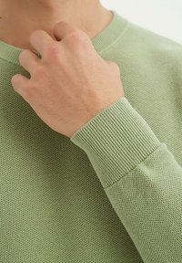 Pull en maille vert clair avec un corps texturé et des poignets côtelés. Le tissu a une apparence douce avec un tissage lâche et respirant.
