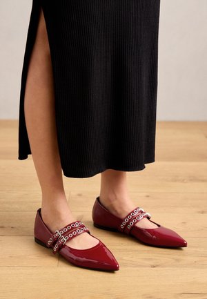Person trägt glänzende bordeauxrote Ballerinas mit spitzer Kappe, doppelten Schnallenriemen und Ösendetails, kombiniert mit einem schwarzen gerippten Rock auf Holzboden.