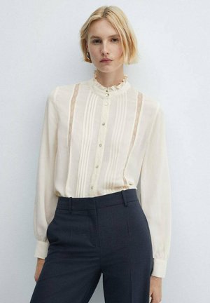 Blonde vrouw in een crèmekleurige blouse met kantdetails en een gerimpelde kraag, gestoken in hoge, donkere, op maat gemaakte broek, tegen een effen achtergrond.