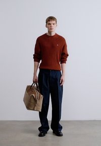 Καφέ ριμπ πλεκτό πουλόβερ, ναυτικό παντελόνι με φαρδιά μπατζάκια, κρατώντας μπεζ σακ βουαγιάζ με καφέ λεπτομέρειες, στέκεται σε ελαφρύ τσιμεντένιο δάπεδο.