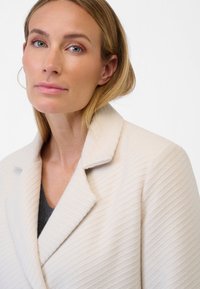 Gelaagd, crèmekleurig blazer met een geknoopte kraag, voorzien van verticale ribbels, gedragen over een donkergrijze top. Eenvoudig en modern ontwerp.