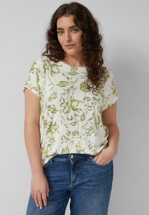 Kobieta o kręconych włosach, ubrana w biały krótki rękaw z zielonym kwiatowym wzorem i niebieskie dżinsy, stoi z jedną ręką w kieszeni na tle szarej ściany.