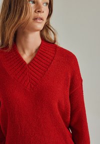 Czerwony sweter o fakturowanej strukturze z głębokim dekoltem w literę V i prążkowanymi akcentami przy kołnierzu. Miękki, puszysty materiał w luźnym kroju.