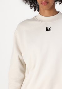 Sudadera beige claro con cuello de canalé y logo negro "HUGO" en el pecho. Textura suave, corte relajado y sin patrones ni acentos visibles.