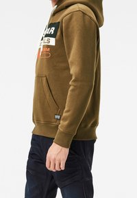 Sudadera color verde oliva con un bolsillo frontal, cordones blancos y detalles del logo impresos en negro y naranja en el pecho. Tejido de mezcla de algodón.