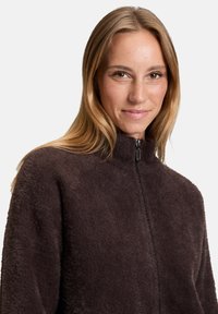 Giacca in pile marrone con zip e colletto alto, caratterizzata da una texture morbida e dettagli di cucitura minimi. La chiusura a zip è visibile.