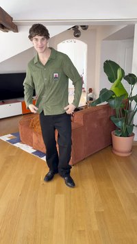Grön manchesterkjorta med knappar och bröstficka, i kombination med svarta byxor och svarta skor, står på trägolv.