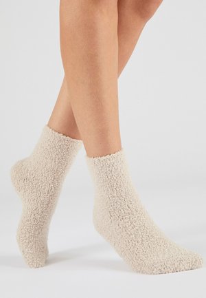 Personne portant des chaussettes de cheville douces, beiges et duveteuses, debout sur une surface gris clair avec les jambes croisées au niveau des mollets.