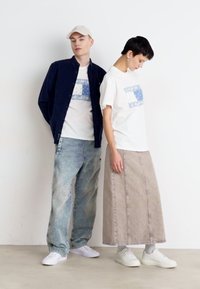 Dos personas están de pie contra un fondo simple. Una lleva una chaqueta azul marino de botones, una camiseta gráfica blanca y jeans de un azul claro. La otra lleva una camiseta blanca, una falda de mezclilla beige y zapatillas blancas.