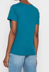 Camiseta corta de manga corta en color verde azulado, hecha de algodón, con un escote redondo y un bajo ligeramente curvado, combinada con pantalones vaqueros de color azul claro.