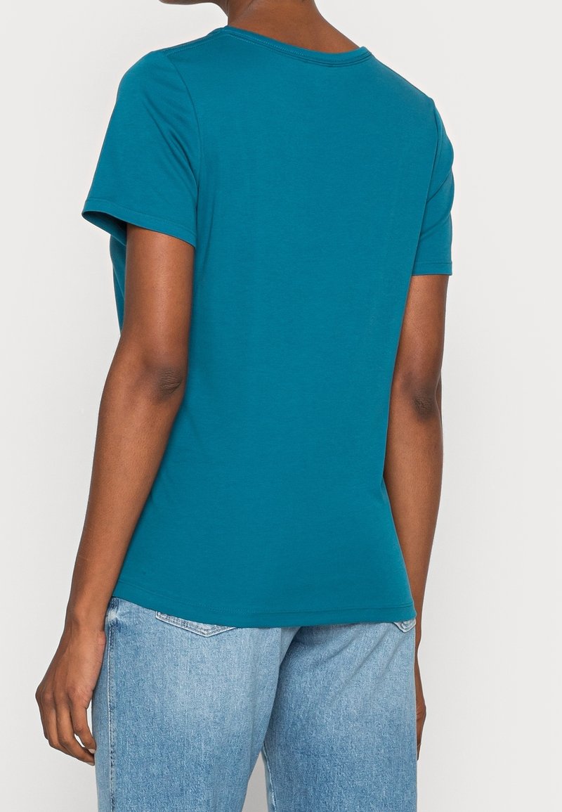 Camiseta corta de manga corta en color verde azulado, hecha de algodón, con un escote redondo y un bajo ligeramente curvado, combinada con pantalones vaqueros de color azul claro.