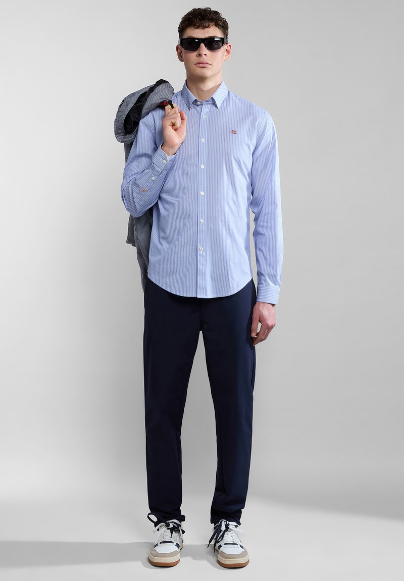 Niebieska i biała koszula w paski na guziki, ciemne spodnie, casualowe sneakersy z białymi, granatowymi i beżowymi akcentami; model trzyma kurtkę, nosi okulary przeciwsłoneczne.