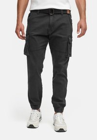Homme portant un pantalon cargo noir avec poches latérales, ceinture noire, baskets blanches et un t-shirt blanc visible à la taille.