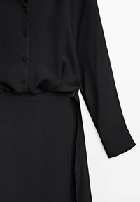 Robe noire à manches longues boutonnée avec une ceinture en tissu assortie autour de la taille sur fond blanc.