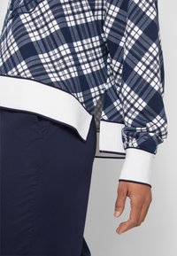 Top a quadri blu navy e bianco con maniche lunghe e polsini bianchi a coste. Presenta una fessura laterale e una texture morbida. Abbinato a pantaloni blu navy.