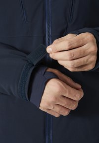 Námořnická modrá bunda se zipem a úpravou manžet na suchý zip; hladká tkanina s přiléhavým střihem, ukazující ruce upravující manžetu.