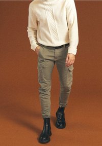 Osoba ubrana w kremowy sweter w sploty, oliwkowe spodnie cargo i czarne skórzane buty, idąca na tle jednolitego brązowego koloru.