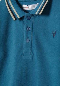 Modra otroška polo majica s tremi gumbi, črtastim ovratnikom, majhnim izvezenim temnim jelenovim logotipom na prsih, vidno označbo velikosti.