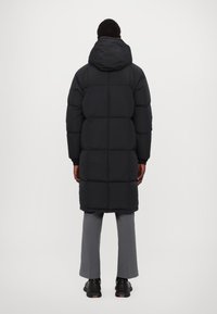 Abrigo de plumas negro con capucha, con diseño acolchado y corte hasta las rodillas, combinado con pantalones grises y zapatillas negras.