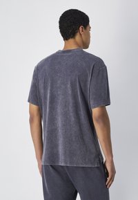 Camiseta gris de gran tamaño con acabado desgastado; mangas cortas, cuello redondo, textura suave, aberturas laterales en el dobladillo, ajuste casual.