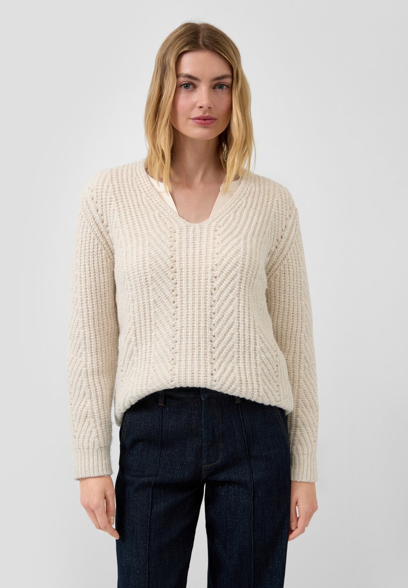 Cremfarbener gestrickter Pullover mit V-Ausschnitt, strukturiertem Muster und langen Ärmeln, kombiniert mit dunklen Jeanshose.