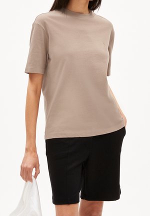 Person iført en beige T-shirt med rund hals og korte ærmer samt sorte knælange shorts, der holder et hvidt klæde i venstre hånd.