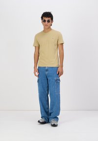 Un tricou din bumbac bej, cu un logo mic, asortat cu pantaloni cargo din denim albastru lărgi, cu buzunare laterale. Purtați cu adidași gri și ochelari de soare.