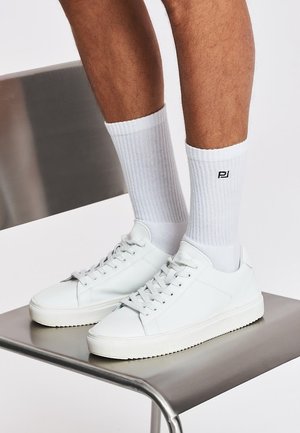 Nogi w białych prążkowanych skarpetkach i białych sneakersach sznurowanych stojące na nowoczesnym metalowym krześle na jednolitym tle.