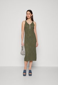 Robe en tricot verte olive avec un décolleté en V, un motif découpé et un ourlet festonné. Associée à des chaussures à plateforme bleues et un petit sac à main à motifs.