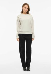 Mujer de pie vistiendo un sudadera beige con el texto "MINIMALISM", pantalones negros y botas de tobillo negras sobre un fondo blanco.