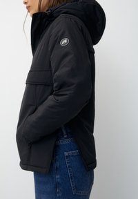 Sort, polstret jakke med hætte, der har en frontlomme og et logo på ærmet. Sættes sammen med blå denim jeans.